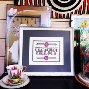 May include: A framed cross-stitch with a white background and black lettering that reads "Feminist Killjoy". The lettering is surrounded by a black border with pink flowers at the top and bottom.
