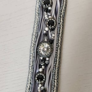 Op de afbeelding: Een grijs en zilveren kralenarmband met een rechthoekig ontwerp. De armband heeft een centraal lintontwerp met zwarte en heldere edelstenen en een zilveren sluiting. De randen zijn afgezet met kleine zilveren kralen.