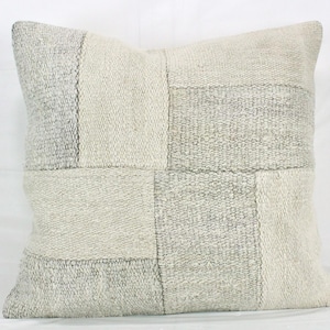 May include: Square decorative pillow with a patchwork design. The pillow features a neutral color palette with various shades of gray and off-white. The textured fabric is divided into rectangular sections.