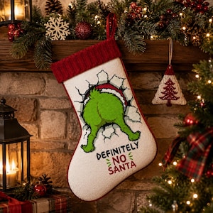May include: A cream-colored Christmas stocking with a red knitted cuff and a hanging loop. The stocking features a needlepoint design of a green character's backside emerging from a hole in a wall, with the text "DEFINITELY NO SANTA" below.