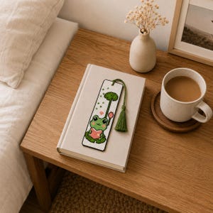 Puede incluir: Marcapáginas hecho a mano con un diseño de rana bordada a punto de cruz sosteniendo un corazón. La rana es verde y rosa, con una borla verde. El marcapáginas está sobre un libro cerrado en una mesa de madera, junto a una taza de café.