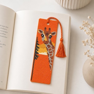 May include: Orange bookmark with a cross-stitched giraffe design. The bookmark features a giraffe's head and neck against a sunset background. A matching orange tassel hangs from the top. The bookmark is resting on an open book.