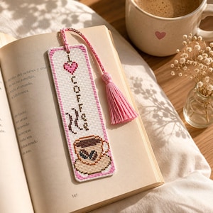 Puede incluir: Un marcapáginas hecho a mano con temática de café. El marcapáginas blanco presenta un borde rosa y un diseño de punto de cruz con una taza de café, vapor y la palabra "COFFEE". Una borla rosa cuelga de la parte superior. Una taza de café y un libro están en el fondo.