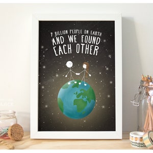 Può includere: Una stampa incorniciata bianca con sfondo nero e un globo blu e verde. Due figure stilizzate sono in piedi sul globo tenendosi per mano. Il testo sulla stampa dice "7 miliardi di persone sulla terra e ci siamo trovati."