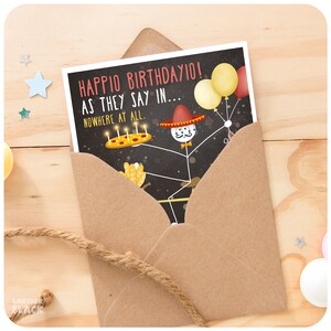May include: A birthday card with a stick figure wearing a sombrero and holding a pizza with candles. The card says "Happio Birthdayio! As they say in... Nowhere at all."