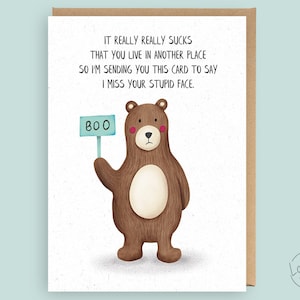 May include: A humorous greeting card featuring a brown bear holding a sign that says "BOO." The card includes the text: "IT REALLY REALLY SUCKS THAT YOU LIVE IN ANOTHER PLACE SO I'M SENDING YOU THIS CARD TO SAY I MISS YOUR STUPID FACE."