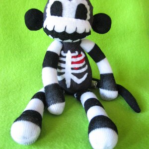 May include: A plush monkey toy crafted from black and white striped fabric. The face features a white skull pattern with black eye sockets and a wide grin. The torso displays a white rib cage graphic with a small, bright red heart detail.