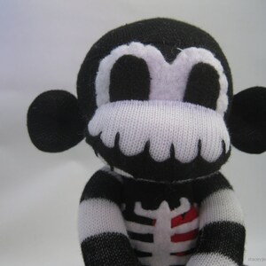 May include: A black and white plush monkey toy made from knit fabric. The monkey features a white skeleton rib cage design on its chest with a red heart detail. It has large round ears, white eye patches, and a white mouth area. The text staceyjean.com is visible.