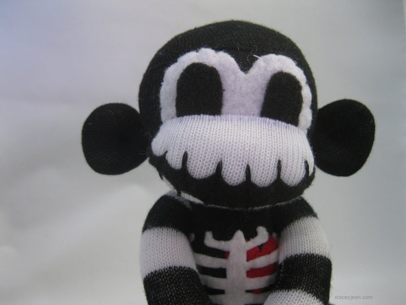 May include: A black and white plush monkey toy made from knit fabric. The monkey features a white skeleton rib cage design on its chest with a red heart detail. It has large round ears, white eye patches, and a white mouth area. The text staceyjean.com is visible.