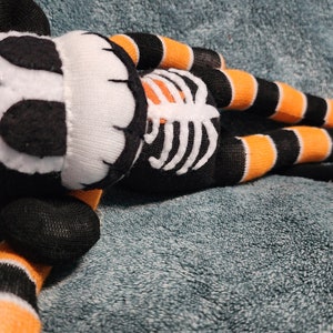 May include: A black and orange striped sock monkey with a white skeleton design on its chest. The monkey is lying on a blue fuzzy surface.