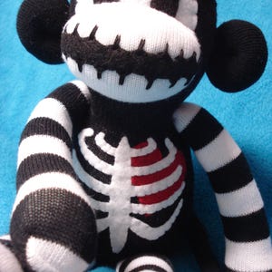 May include: A plush monkey toy made from black and white knit fabric. The monkey features a skeleton rib cage design on its chest with a red heart accent, striped arms and legs, and a white face mask with black stitched details.