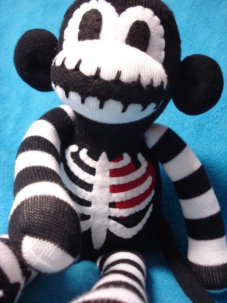 May include: A plush monkey toy made from black and white knit fabric. The monkey features a skeleton rib cage design on its chest with a red heart accent, striped arms and legs, and a white face mask with black stitched details.