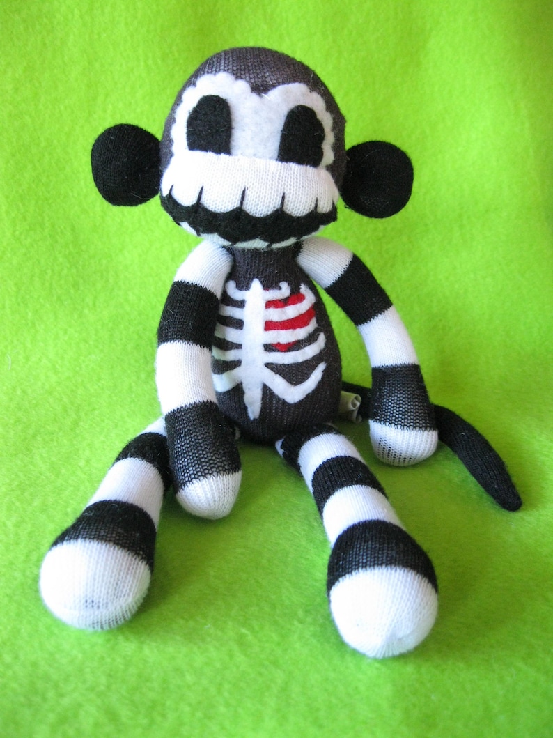 May include: A plush monkey toy crafted from black and white striped fabric. The face features a white skull pattern with black eye sockets and a wide grin. The torso displays a white rib cage graphic with a small, bright red heart detail.