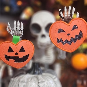 May include: Two orange heart-shaped Halloween keychains with jack-o'-lantern faces. Each keychain has a skeleton hand holding a silver ring. One has a green strap, the other a brown strap. The background includes a skeleton and orange decorations.