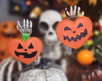 May include: Two orange heart-shaped Halloween keychains with jack-o'-lantern faces. Each keychain has a skeleton hand holding a silver ring. One has a green strap, the other a brown strap. The background includes a skeleton and orange decorations.