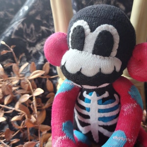 May include: A black and white sock monkey with a skeleton design. The monkey has pink ears and blue and white polka dot arms and legs.