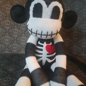 May include: A plush monkey doll crafted from black and white knit fabric. The doll features a stitched mouth, a white ribcage graphic on the chest, and a small red heart patch. It has large round ears and a striped pattern on its limbs.