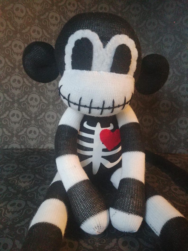 May include: A plush monkey doll crafted from black and white knit fabric. The doll features a stitched mouth, a white ribcage graphic on the chest, and a small red heart patch. It has large round ears and a striped pattern on its limbs.