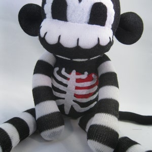 May include: A plush monkey toy made from black and white striped fabric. The monkey features a white skeleton rib cage applique on its chest with a red heart, and a white face mask with large black eyes and a wide, toothy grin.