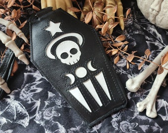 May include: Black coffin-shaped wallet with a white skull and moon design. The wallet has a star at the top and three vertical lines at the bottom. Decorative elements include a skeleton hand, bones, and a floral pattern.