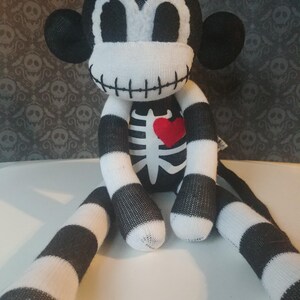 May include: A plush monkey toy made of black and white striped fabric. The monkey features a white face with large black eyes, a stitched mouth, and a white skeleton rib cage design on its chest with a small, bright red heart appliqu&eacute;.