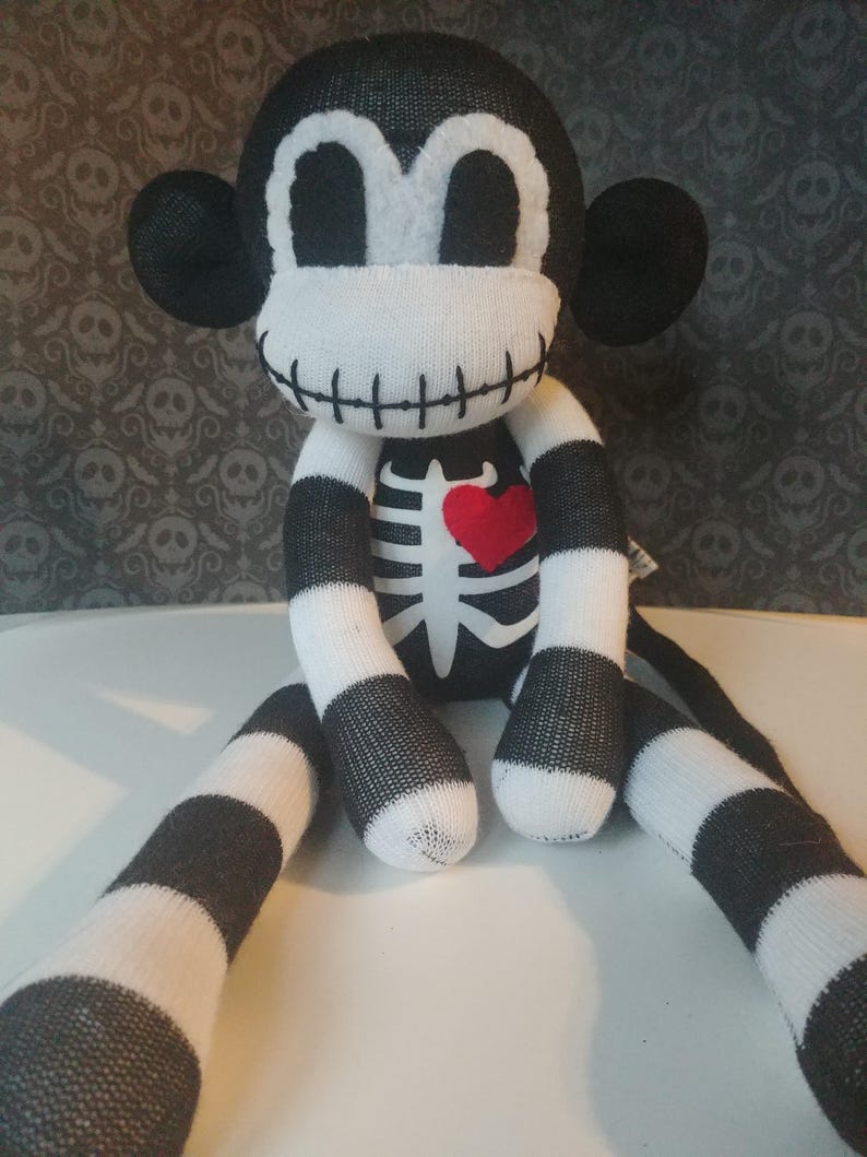 May include: A plush monkey toy made of black and white striped fabric. The monkey features a white face with large black eyes, a stitched mouth, and a white skeleton rib cage design on its chest with a small, bright red heart appliqu&eacute;.