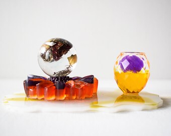 Può includere: Una scultura artistica in resina. Un globo trasparente con elementi incorporati poggia su una base a forma di fiore, con segmenti arancioni e viola. È presente anche un bicchiere sfaccettato, giallo e viola.
