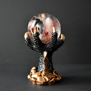 Op de afbeelding: Een zwart drakenklauw beeldje dat een heldere glazen bol vasthoudt met rode en witte accenten. De basis van het beeldje is goud.