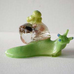 May include: A green glass snail sculpture with a clear glass shell filled with colorful inclusions. The shell is topped with a cluster of green glass buds.