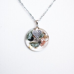 Op de afbeelding: Een zilveren ketting met een ronde hanger met groene en bruine stenen, koperdraad en een zilveren hartbedel.