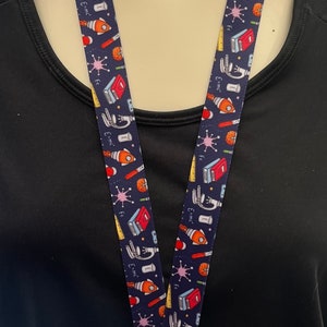 May include: A blue lanyard with a white background and colorful science-themed illustrations, including a rocket, microscope, and books. The lanyard has a silver clip at the end.