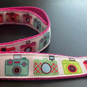 May include: A pink and white ribbon with a silver metal buckle. The ribbon features a repeating pattern of colourful cameras.