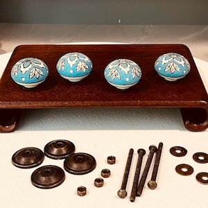 May include: Four round ceramic knobs featuring a bright blue and white floral pattern with dark outlines. The set includes four dark metal circular backplates, four long threaded bolts, four small metal nuts, and four flat metal washers.