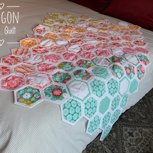 May include: A colorful baby quilt made of hexagon-shaped fabric pieces. The quilt is made in a rainbow pattern with white, pink, green, yellow, and orange fabrics. The quilt is laid out on a white bed with a striped pattern.