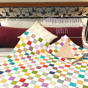 May include: Twin checkerboard quilts in a bedroom setting. The quilts feature a colorful patchwork design with squares of various colors and patterns. The text "TWIN CHECKERBOARD QUILTS" is written in white above the quilts.