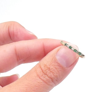 May include: A delicate gold ring featuring a row of small, vibrant green gemstones and clear stones. The ring is held between fingers against a white background, showcasing its slender design and sparkling details. Jewelry.