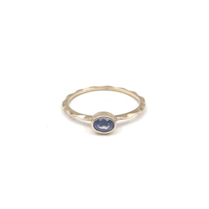 Op de afbeelding: Een gouden ring met een golvende band en een ovale, blauwe edelsteen in het midden. De ring wordt gepresenteerd tegen een witte achtergrond, waardoor het ontwerp en de kleur van het sieraad worden benadrukt.