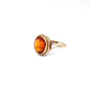 May include: A gold ring featuring a large, round, faceted orange gemstone set in a bezel with a decorative twisted rope border. The band is made of polished gold and splits into two thin, elegant arms where it connects to the central setting.