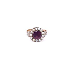 May include: A gold ring featuring a central round deep purple gemstone surrounded by a halo of small white pearls. The band is thin and metallic, supporting the circular cluster setting that holds the dark purple stone and the surrounding pearls.