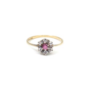May include: A gold ring with a central pink gemstone, encircled by smaller, clear stones. The ring has a simple gold band, and the gemstones are set in a silver-toned metal. The design is floral-inspired.