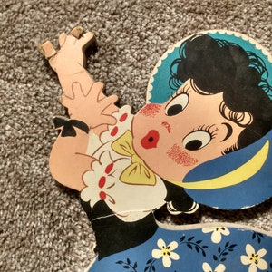 Peut inclure: D&eacute;coupe en bois vintage et color&eacute;e d'une fillette de dessin anim&eacute; aux cheveux noirs, avec un chapeau bleu et une robe bleue &agrave; fleurs blanches. La fille a des joues roses et une expression surprise. La d&eacute;coupe est sur un tapis gris.