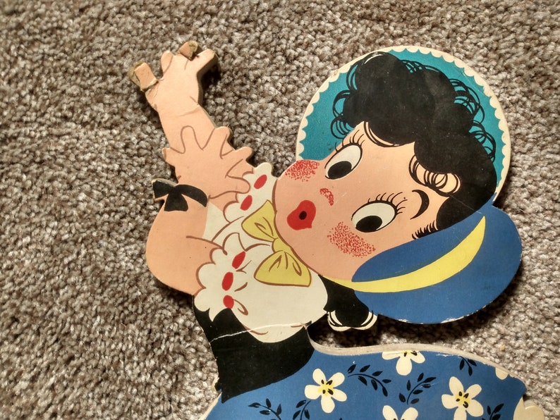 Peut inclure: D&eacute;coupe en bois vintage et color&eacute;e d'une fillette de dessin anim&eacute; aux cheveux noirs, avec un chapeau bleu et une robe bleue &agrave; fleurs blanches. La fille a des joues roses et une expression surprise. La d&eacute;coupe est sur un tapis gris.