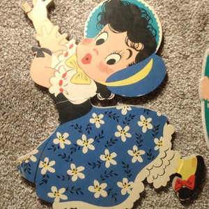 Peut inclure: D&eacute;coupe en bois vintage d'une fille dans une robe bleue avec des motifs floraux blancs. La fille a les cheveux noirs, un chapeau bleu et un n&oelig;ud jaune. La d&eacute;coupe est peinte avec des couleurs vives et a un design fantaisiste, rappelant un jouet pour enfants.