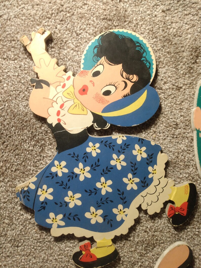Peut inclure: D&eacute;coupe en bois vintage d'une fille dans une robe bleue avec des motifs floraux blancs. La fille a les cheveux noirs, un chapeau bleu et un n&oelig;ud jaune. La d&eacute;coupe est peinte avec des couleurs vives et a un design fantaisiste, rappelant un jouet pour enfants.