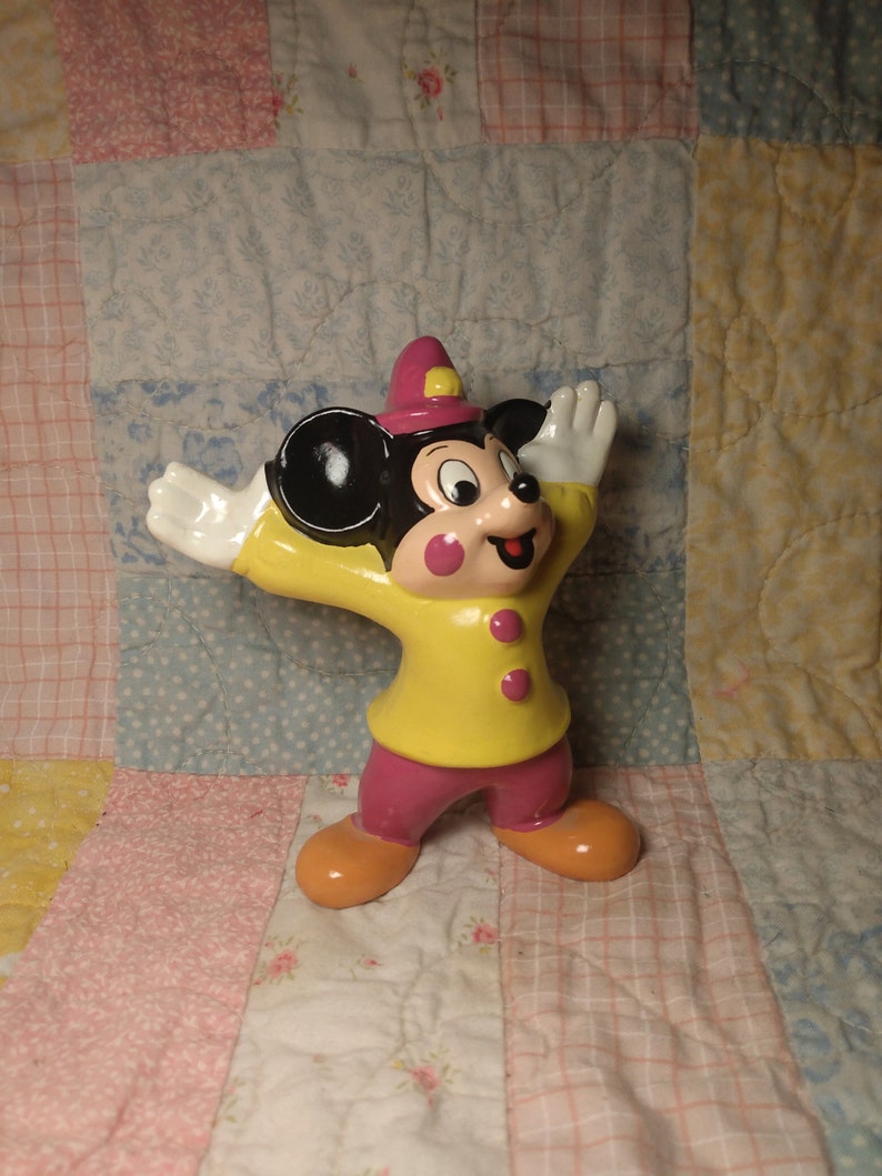 Puede incluir: Una figurita de cer&aacute;mica de Mickey Mouse con un atuendo amarillo y rosa con un sombrero morado. Est&aacute; de pie con los brazos en alto y tiene una gran sonrisa.
