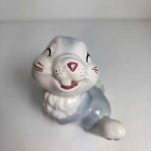 May include: A small ceramic rabbit figurine featuring a glossy white and soft grey finish. The rabbit has a painted pink heart-shaped nose, dark brown whiskers, and closed eyes. It is depicted in a seated position with a textured white chest.