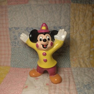Puede incluir: Una figurita de cer&aacute;mica amarilla y rosa de Mickey Mouse con un sombrero rojo y una chaqueta amarilla. Est&aacute; de pie con los brazos extendidos y tiene una gran sonrisa.