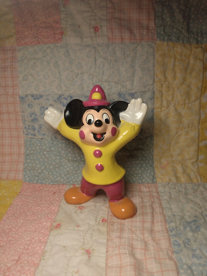 Puede incluir: Una figurita de cer&aacute;mica amarilla y rosa de Mickey Mouse con un sombrero rojo y una chaqueta amarilla. Est&aacute; de pie con los brazos extendidos y tiene una gran sonrisa.