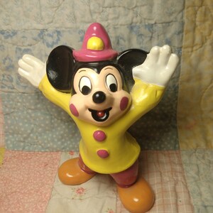 Puede incluir: Una figurita de cer&aacute;mica amarilla y rosa de Mickey Mouse con un sombrero rosa con un bot&oacute;n amarillo. Mickey Mouse est&aacute; de pie con los brazos extendidos y tiene una gran sonrisa en la cara.