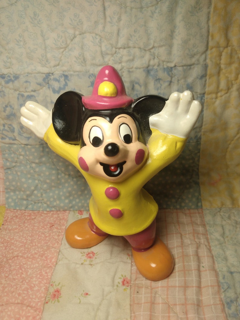 Puede incluir: Una figurita de cer&aacute;mica amarilla y rosa de Mickey Mouse con un sombrero rosa con un bot&oacute;n amarillo. Mickey Mouse est&aacute; de pie con los brazos extendidos y tiene una gran sonrisa en la cara.